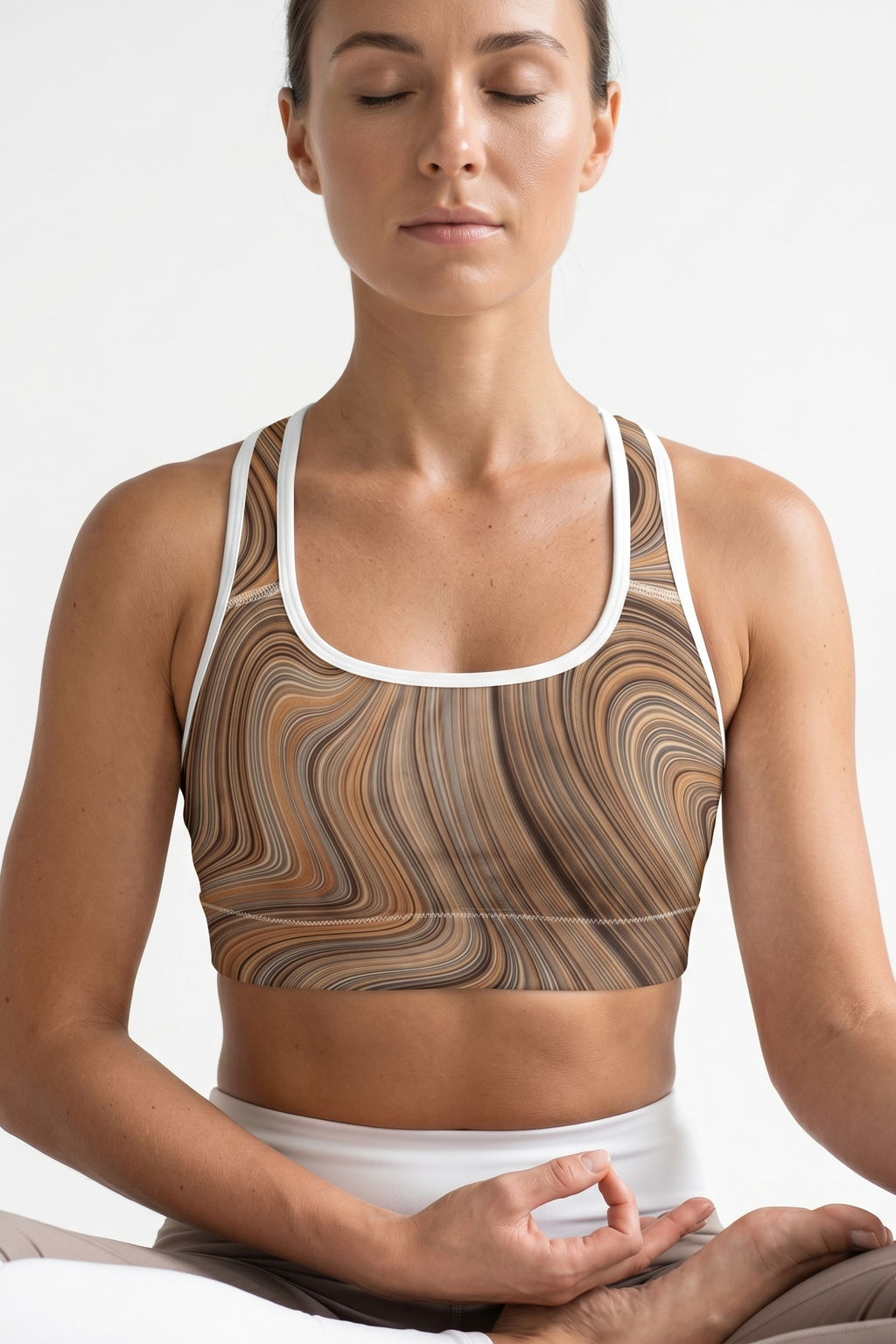 Woman wearing a patterned sports bra and white pants, sitting in a meditative pose on a white background.
