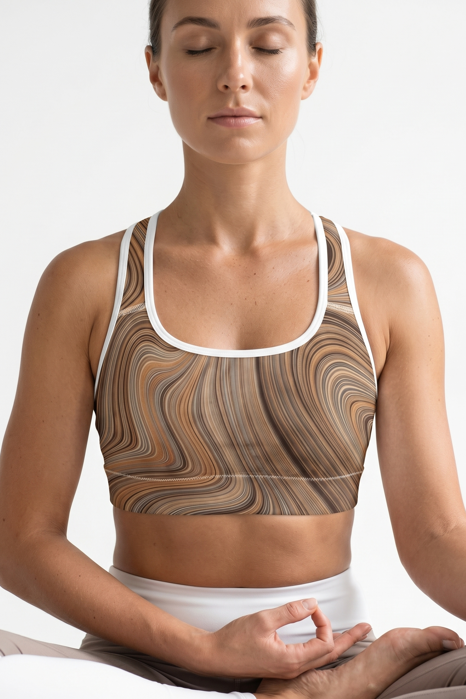 Woman wearing a patterned sports bra and white pants, sitting in a meditative pose on a white background.